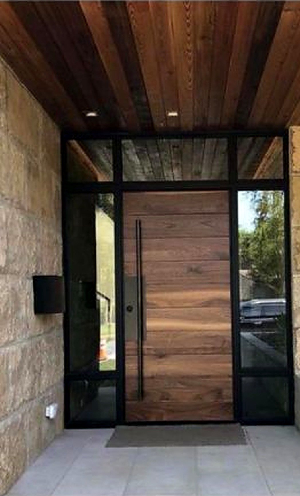 Hardwood pivot entry door in a black steel-framed glass entry beneath a wood ceiling
